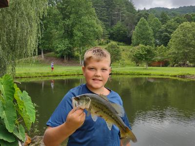 Young boy holding largemouth bass beside pond with trees and cloudy sky