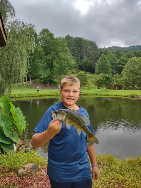 Young boy holding largemouth bass beside pond with trees and cloudy sky