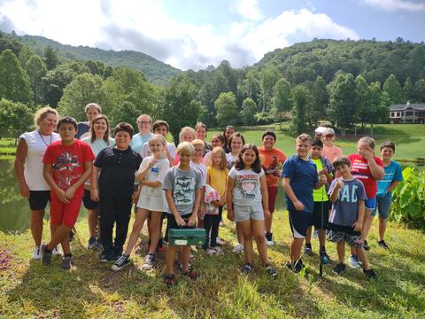 Group of children and adults standing on grass by a lake with tree-covered hills in background