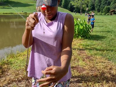 Child wearing "MOSSY OAK" cap holding a fishing rod and small fish by a pond.