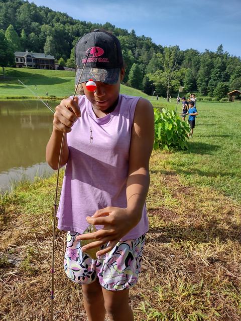 Child wearing "MOSSY OAK" cap holding a fishing rod and small fish by a pond.