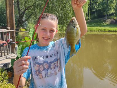 Girl holding a fishing rod and a green sunfish by a pond, wearing a "FRIENDS" shirt