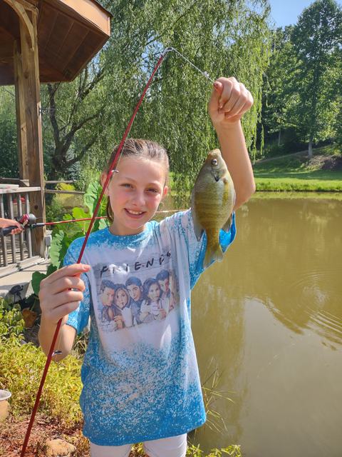 Girl holding a fishing rod and a green sunfish by a pond, wearing a "FRIENDS" shirt