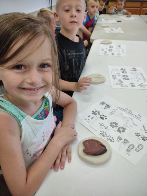 Young girl at table with clay impression and animal track guide, other children seated nearby