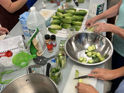 People slicing cucumbers and filling jars for pickling; bottle labeled "DISTILLED WHITE VINEGAR"