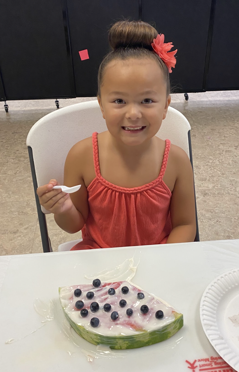 Girl in red dress holding a spoon, yogurt-covered watermelon slice topped with blueberries.