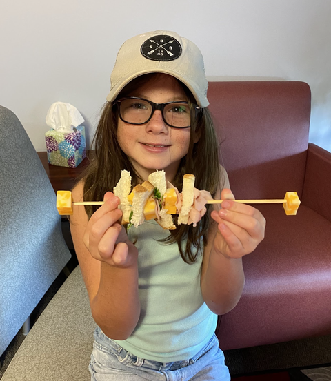 Girl wearing cap and glasses holding a skewer threaded with bread, cheese, and ham