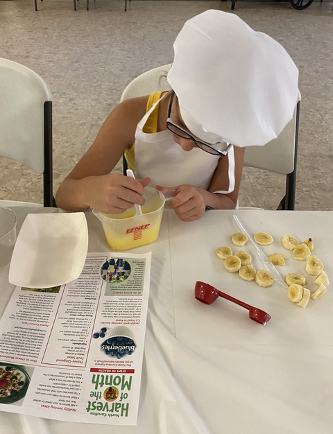 Child in chef hat stirring yellow mixture, EFNEP container and "Harvest of the Month" leaflet