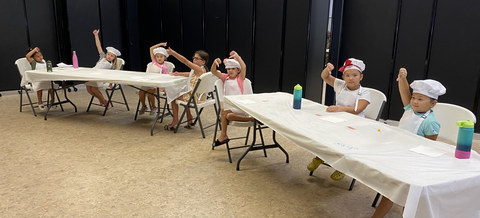 Seven children in chef hats and aprons seated at tables, raising their hands