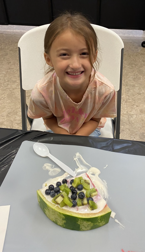 Young girl seated behind a watermelon slice topped with yogurt, diced kiwi, and blueberries