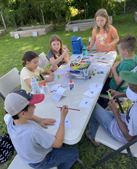 Children seated at a folding table outdoors drawing, coloring, and using glue.
