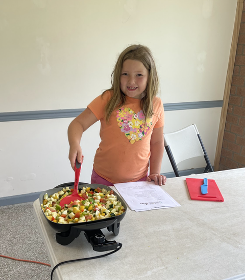 Young girl stirring diced vegetables in an electric skillet on a table with a recipe.