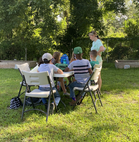 Group of children seated at outdoor table with an adult standing nearby