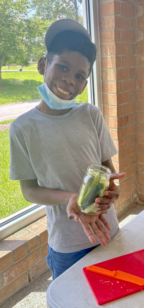 Young boy with mask under chin holding a jar of pickles by a window