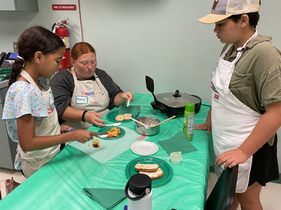 Three children in aprons preparing food at a table; visible "FIRE EXTINGUISHER" sign and name tag "Makayla"
