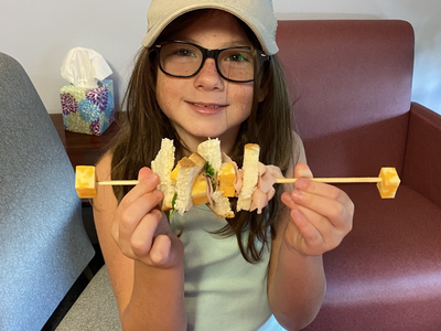 Girl wearing cap and glasses holding a skewer threaded with bread, cheese, and ham