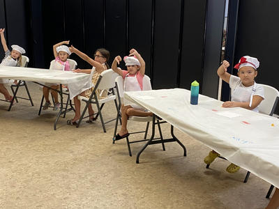 Seven children in chef hats and aprons seated at tables, raising their hands