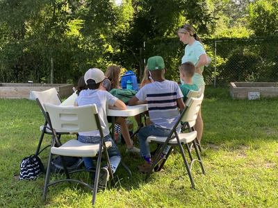 Group of children seated at outdoor table with an adult standing nearby
