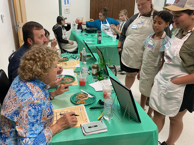 Children in aprons presenting food to seated judges at a green-covered table
