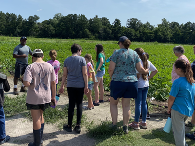 Group of adults and children standing at the edge of a crop field listening to a man speaking
