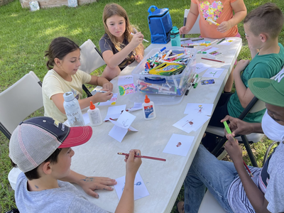 Children seated at a folding table outdoors drawing, coloring, and using glue.