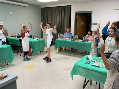 Kids in aprons standing around green-covered tables with hands raised