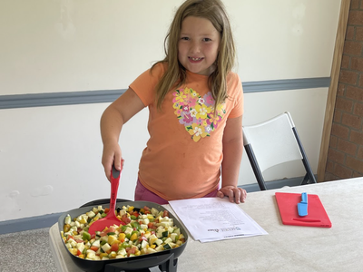 Young girl stirring diced vegetables in an electric skillet on a table with a recipe.