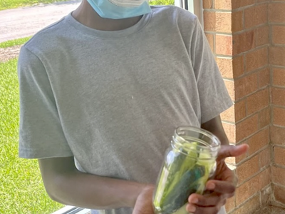 Young boy with mask under chin holding a jar of pickles by a window