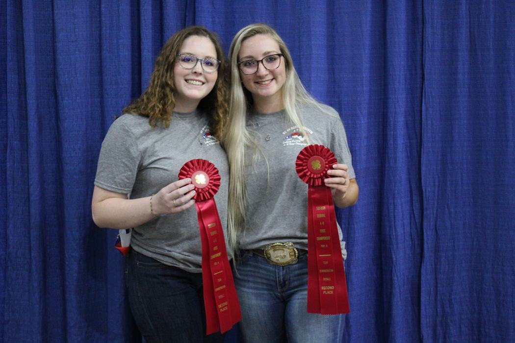 Two women in front of blue curtain holding red rosette ribbons labeled SECOND PLACE