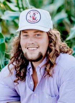 young person with long hair wearing a white cap labeled "NC STATE" and a light purple shirt