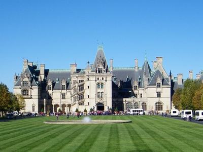 A large white walled estate sits behind a large lawn and fountain. Tourists move towards the front door of the home.