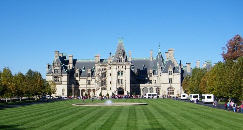 A large white walled estate sits behind a large lawn and fountain. Tourists move towards the front door of the home.