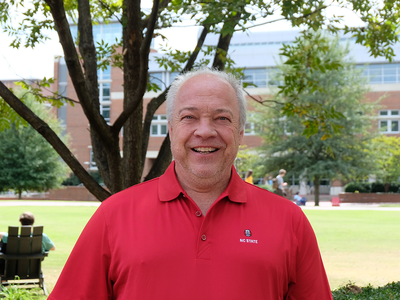 Man wearing red "NC STATE" polo standing on campus lawn under a tree