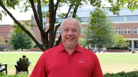Man wearing red "NC STATE" polo standing on campus lawn under a tree