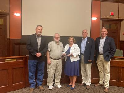 Five people standing in a council chamber; woman holding a glass award; wall sign "IN GOD WE TRUST"
