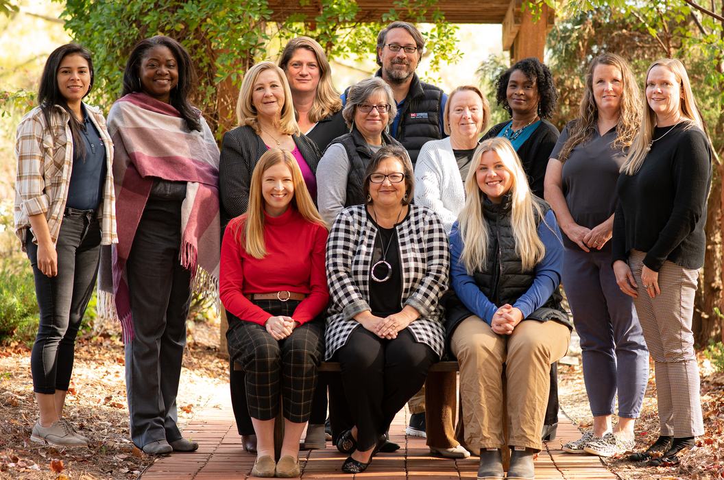 Thirteen adults posed in two rows on a wooden path under trees