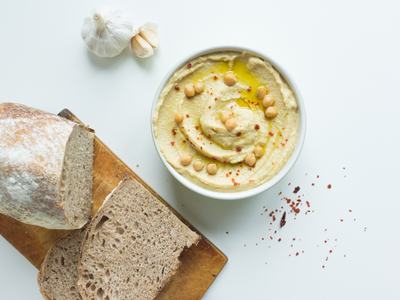 Bowl of hummus with chickpeas, olive oil and chili flakes beside sliced bread and garlic