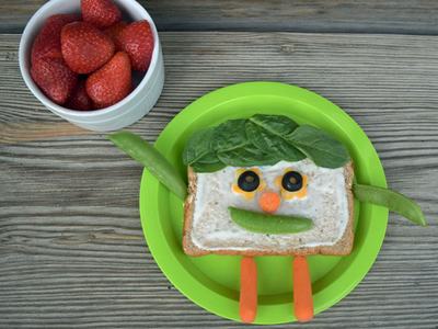 A sandwich has vegetable face and vegetables with a face on plate, strawberries in bowl.