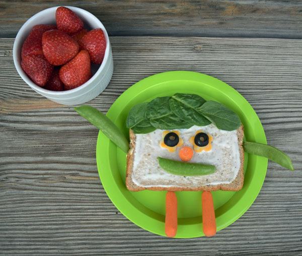 A sandwich made with fresh vegetables for the face on plate, strawberries in bowl.