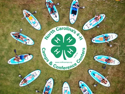 People on paddleboards in a circle around 4‑H logo: "North Carolina's 4‑H Camps & Conference Centers"
