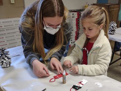 Adult wearing mask helps child assemble holiday craft on table