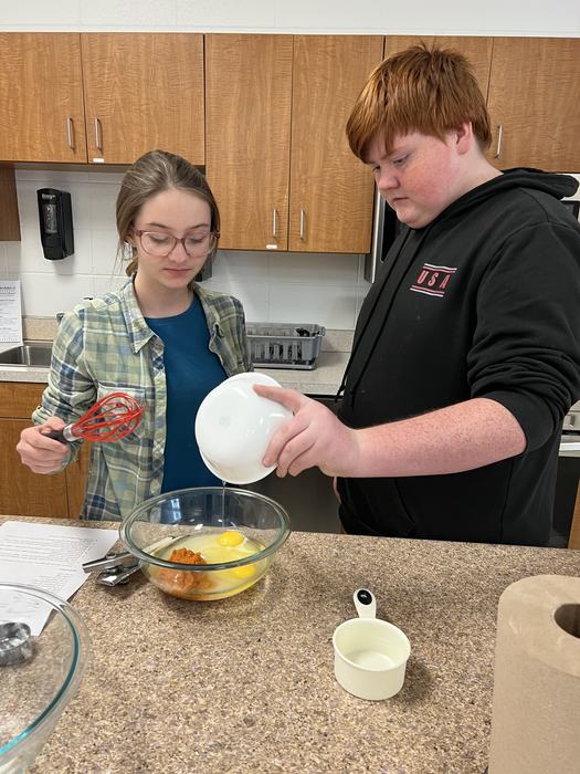 Two students add wet ingredients for pumpkin bread to a mixing bowl.