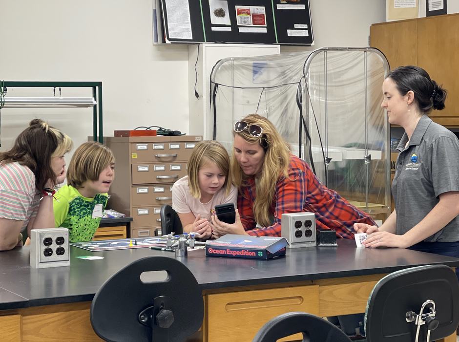 Volunteers and participants gather around an ocean-themed board game while an augmented reality video clip plays. The board game included ocean trivia and shared about challenges scientists encounter when studying it.