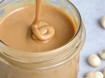 Jar of creamy peanut butter with spoon drizzling into it, shelled peanuts nearby