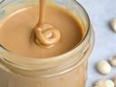 Jar of creamy peanut butter with spoon drizzling into it, shelled peanuts nearby