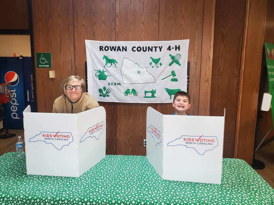 Two kids behind "Kids Voting North Carolina" voting booths at a Rowan County 4‑H table