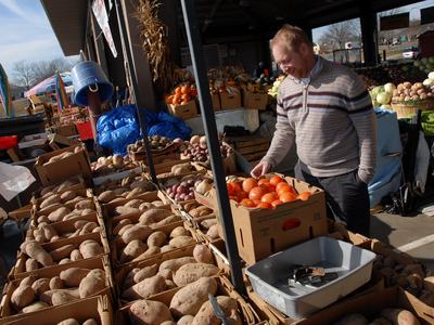 Man selecting tomatoes at an outdoor produce market stall with boxes of potatoes.