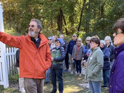 Man in orange jacket gestures while a group listens outdoors near trees and white picket fence