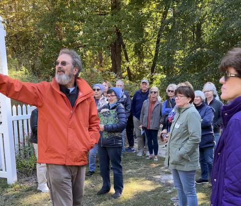 Man in orange jacket gestures while a group listens outdoors near trees and white picket fence