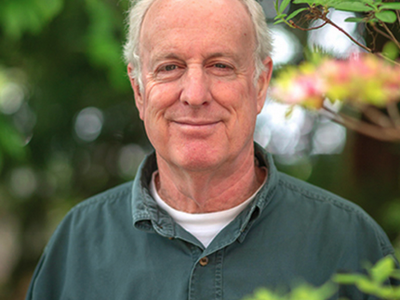 Older man standing outdoors wearing a green shirt, blurred foliage and pink flowers nearby